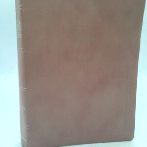 May include: A hardcover book with a muted brown cover. The spine features the title "The Bible" in white lettering, along with author information. The book's edges are slightly worn, suggesting age and use.