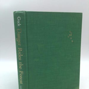 May include: A hardcover book titled "Guck Danger Rides the Forest" by Vanguard. The book has a textured, dark green cover with gold lettering on the spine. The book's pages are visible on the right side.