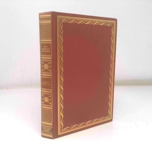May include: A hardcover book with a red cover and gold decorative trim. The spine is a lighter brown with gold lettering and decorative patterns. The book is standing upright against a white background.