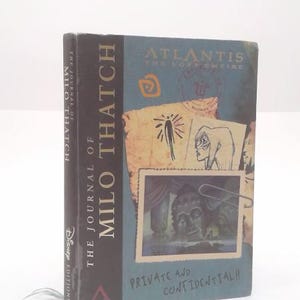 May include: A hardcover book titled "The Journal of Milo Thatch" from Disney Editions. The cover features the title "Atlantis The Lost Empire" with illustrations and the text "Private and Confidential!!". The spine is dark with the title.