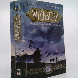 May include: A paperback book cover with a dark blue and purple background featuring a stormy sky and a woman's eyes. The title "Witch Storm" is in gold lettering on a brown banner. The subtitle is "Immortal Magic - Infinite Vengeance". The author's name, "James Clemens", is printed in white lettering on the spine.