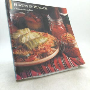 May include: A cookbook titled "Flavors of Hungary" by Charlotte Slovak Biro. The cover features a vibrant image of Hungarian cuisine, including stuffed cabbage rolls and preserved vegetables in glass jars. The book is angled on a white surface.