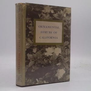 May include: A vintage book titled "Ornamental Shrubs of California." The cover features a floral pattern in shades of green and brown, with a rectangular label in the center displaying the title. The spine has the title printed vertically.