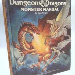 May include: A hardcover book titled Official Advanced Dungeons & Dragons Monster Manual by Gary Gygax. The cover features a large red dragon breathing fire, surrounded by winged horses against a dark blue background. The spine is tan and black.