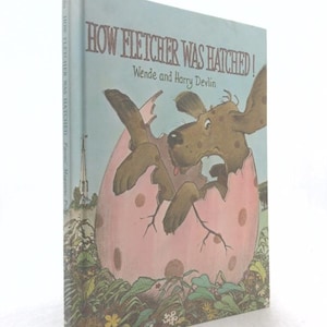 May include: A hardcover children's book titled How Fletcher Was Hatched! by Wende and Harry Devlin. The cover features a brown dog emerging from a large, cracked, pink polka-dot egg, set against a light blue sky and green grassy foreground.