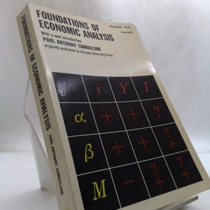 May include: A hardcover book titled "FOUNDATIONS OF ECONOMIC ANALYSIS" by Paul Anthony Samuelson. The cover features a black grid with mathematical symbols in yellow and red. The book is propped up on a clear stand.