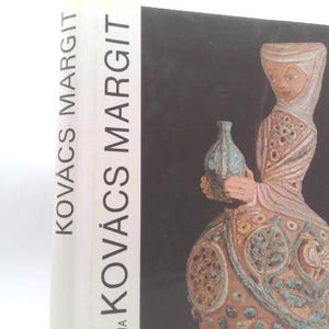 May include: A book titled "Kovács Margit" stands upright, next to a black background. The book's spine displays the title in large, bold letters. The right side of the book features a ceramic sculpture of a woman holding a vase, with intricate details and a muted color palette.
