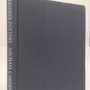 May include: A hardcover book titled "Pioneer Pottery" by Michael Cardew. The book has a dark blue cover with the title and author's name in silver lettering on the spine. The book is standing upright.