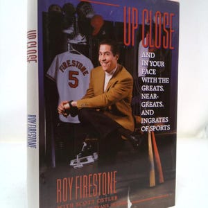 May include: A book titled "Up Close and In Your Face With The Greats, Near-Greats, And Ingrates Of Sports" by Roy Firestone with Scott Ostler. The book cover features a photo of Roy Firestone sitting in a locker room with a baseball jersey that says "Firestone 5" on it.