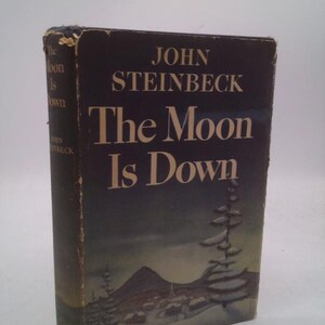 May include: A vintage hardcover book titled "The Moon Is Down" by John Steinbeck. The cover is dark with white lettering and a winter landscape illustration. The book shows signs of wear, with a slightly distressed appearance.