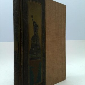 May include: A vintage hardcover book with a brown linen cover and a spine featuring a gold illustration of the Statue of Liberty. The title of the book is "The Real America in Pictures".