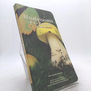 May include: A book titled "Mushrooms of Colorado and the Southern Rocky Mountains" is displayed. The cover features a large mushroom with a yellow cap and white stem against a green background. The author's name and publishing information are also visible.