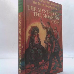 May include: A vintage hardcover book titled "The Mystery of the Moaning Cave" by Alfred Hitchcock. The cover features an illustration of three investigators and a skeleton. The book is part of the Alfred Hitchcock Mystery Series.