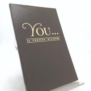 May include: A brown book titled "You..." by Frances Wilshire. The title and author's name are in gold lettering. The book is displayed on a clear plastic stand against a white background.