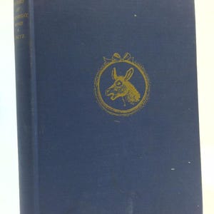 May include: A vintage hardcover book with a navy blue cover. The cover features a gold-colored illustration of a donkey inside a decorative frame. The book's spine displays the title and author's name in gold lettering.