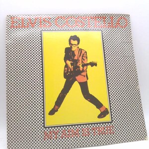 May include: Album cover for "My Aim Is True" by Elvis Costello. The album art features a stylized image of Costello playing a guitar, set against a yellow background. The album cover has a black and white checkered pattern and the text "ELVIS COSTELLO" and "MY AIM IS TRUE".