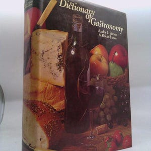 May include: A hardcover book titled "Dictionary of Gastronomy" with a cover illustration featuring cheese, bread, wine, fruit, and a wine glass. The book's spine is black with white text.