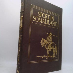 May include: A brown leather-bound book with gold lettering on the spine and front cover. The title is "Sport in Somaliland".