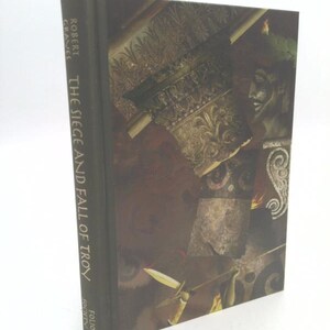 May include: A hardcover book titled "The Siege and Fall of Troy" by Robert Graves. The book's cover features a dark, artistic illustration with architectural elements and a bust. The spine displays the author's name and title.
