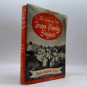 May include: A hardcover book titled The Story of the Trapp Family Singers by Maria Augusta Trapp. The cover features a black and white photograph of a group of people in a field, enclosed in a red oval, with a red spine and white text.