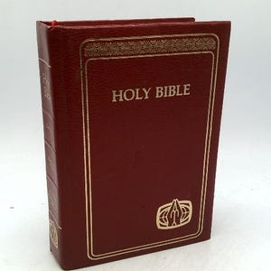 May include: A burgundy-colored Holy Bible with gold lettering and accents. The book has a rectangular shape with the words "HOLY BIBLE" prominently displayed on the cover. A gold emblem is at the bottom of the cover.