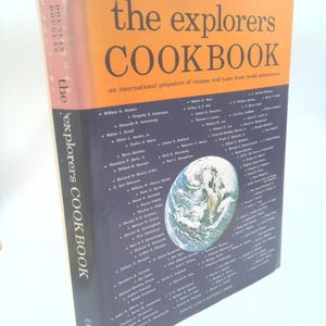 May include: A hardcover book titled "the explorers COOKBOOK" with an orange cover and black text. The book's spine is pink with the author's name, Douglas, repeated. The cover features a world map graphic and a list of names.