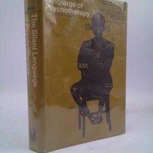 May include: A hardcover book titled "The Silent Language of Psychotherapy" with a mustard yellow cover. The cover features a silhouette of a person sitting on a chair, with symbols and text. The book is wrapped in clear plastic.
