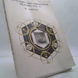 May include: A book titled "GEM ELIXIRS AND VIBRATIONAL HEALING, VOL. II" by Gurudas. The cover is off-white with a black and gold circular design. The center of the design features a grayscale image. The book is slightly angled.