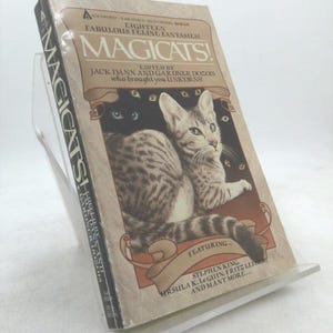 May include: A paperback book titled "MAGICATS!" is displayed on a clear stand. The cover features a tabby cat with other cat eyes in the background. The book's title is in large letters, with the subtitle "Eighteen Fabulous Feline Fantasies!" and the names of the editors and featured authors.
