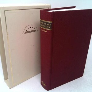 May include: A maroon hardcover book titled "Writings on the Constitution, Part Two" with a gold-lettered spine, next to a beige book sleeve. The book sleeve has the words "The Library of America" and a logo.