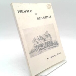 May include: A vintage book titled "Profile of San Dimas" by J. Howard Hoover. The cover is off-white with black text and a black and white illustration of a building. The book is propped up on a clear stand.