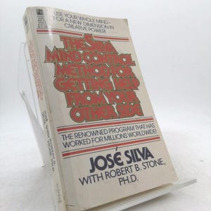 May include: A vintage paperback book titled "The Silva Mind Control Method for Getting Help from Your Other Side" by José Silva and Robert B. Stone, Ph.D. The book has a white cover with red and orange lettering and is displayed on a clear plastic stand.