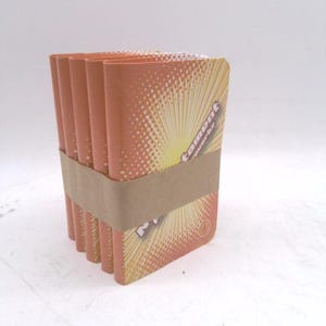 May include: A stack of orange-colored rectangular items, bound together with a brown band. The items have a design featuring a sunburst pattern and the word "tament" in a stylized font. The background is plain white.