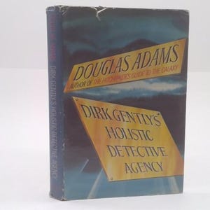 May include: A hardcover book titled "Dirk Gently's Holistic Detective Agency" by Douglas Adams. The cover features a blue and yellow design with the author's name and the book's title prominently displayed. The spine also has the book's title.