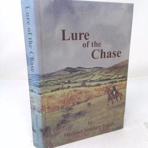May include: A hardcover book titled Lure of the Chase by Michael Sinclair-Smith. The cover features a landscape illustration of a person riding a horse followed by a pack of hounds in a field with rolling hills under a cloudy sky.