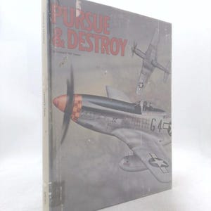 May include: A hardcover book titled Pursue & Destroy by Leonard 'Kit' Carson. The cover features a gray illustration of a P-51 Mustang fighter plane with a red nose and checkered pattern, alongside a second plane flying in the background.