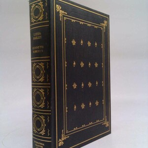 May include: A hardcover book titled "Lydia Bailey" by Kenneth Roberts. The book has a black cover with gold decorative accents and text. The spine features gold circular designs and the title. The book is part of the International Collectors Library.