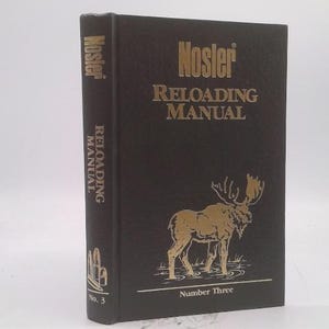 May include: A dark brown "Nosler Reloading Manual" book with gold lettering and a gold moose illustration. The book is labeled "Number Three" at the bottom. The spine reads "Nosler Reloading Manual No. 3".