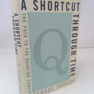 May include: A hardcover book titled "A Shortcut Through Time" by George Johnson. The cover features the title in dark blue, a large "Q" and a background of binary code in light blue. The spine also displays the title and author.