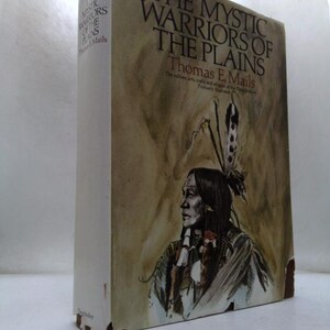 May include: A hardcover book titled The Mystic Warriors of the Plains by Thomas E. Mails. The cover features a painted illustration of a person with long dark hair and a feather headdress against a tan background. The book shows signs of wear.