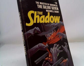 May include: A vintage paperback book titled "The Shadow" by Maxwell Grant. The cover features a dark illustration with the title in yellow, a figure in a hat and red scarf, and hands reaching for two guns. The book is slightly angled.