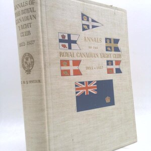 May include: A hardcover book titled "Annals of the Royal Canadian Yacht Club 1852-1957." The cover is beige with various flags and the British Union Jack. The book's spine displays the title and author's name.