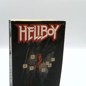 May include: A paperback book titled Hellboy: On Earth As It Is In Hell by Brian Hodge. The black cover features a red-skinned character in a trench coat surrounded by floating symbols, with the title in large red letters at the top.