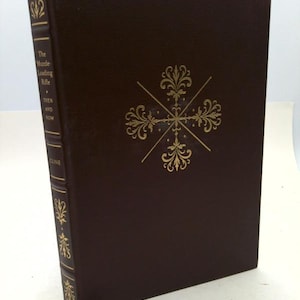 May include: A hardcover book titled "The Muzzle-Loading Rifle Then and Now" with a dark brown cover and gold decorative accents. The spine features gold text and embellishments. A maroon ribbon bookmark is visible.