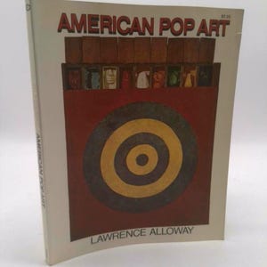 May include: A book titled "AMERICAN POP ART" by Lawrence Alloway. The cover features a red and brown design with a bullseye and portraits. The title is in red letters with a white background.