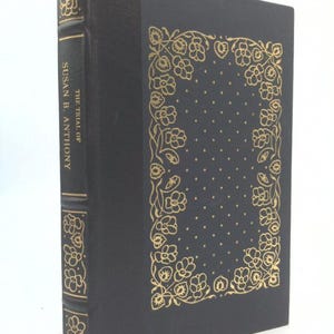 May include: A hardcover book titled "The Trial of Susan B. Anthony." The book has a black cover with gold floral designs and gold lettering on the spine. The cover features a dotted pattern within a floral border.