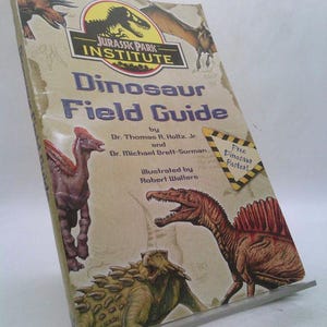 May include: A well-worn "Jurassic Park Institute Dinosaur Field Guide" book. The cover features illustrations of various dinosaurs, including a Spinosaurus and a Pterodactyl. The book's title is in large, blue letters, with the text "Free Dinosaur Poster!" on a yellow sign.