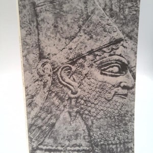 May include: A rectangular book with a black and white cover featuring a detailed stone relief carving of an ancient figure in profile. The figure wears a tall, pointed hat and has a stylized, curly beard and hair. The book spine is visible.