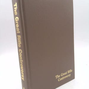 May include: A hardcover book titled "The Great Rifle Controversy" in a brown cover. The title is printed in gold lettering on the spine and front cover. The book is standing upright against a white background.