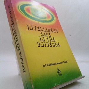 May include: A paperback book titled "Intelligent Life in the Universe" by I.S. Shklovskii and Carl Sagan. The cover features a yellow background with a green and orange design and the title in bold text. The book is propped up on a clear stand.